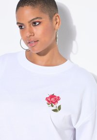 Sudadera blanca con bordado de una rosa rosa y verde en el área del pecho izquierdo, con un cuello redondo y un corte relajado.