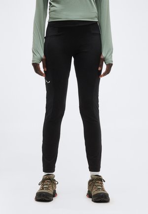 Άτομο που φοράει μαύρο αθλητικό κολάν με υφασμάτινες πλαϊνές πάνελ, πράσινο μακρυμάνικο πάνω μέρος, λευκές κάλτσες και καφέ ορειβατικά παπούτσια, στέκεται όρθιο.