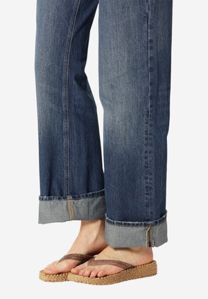 Persoon die opgerolde blauwe spijkerbroeken en bruine gevlochten platform slippers draagt, met voeten en onderbenen zichtbaar tegen een witte achtergrond.