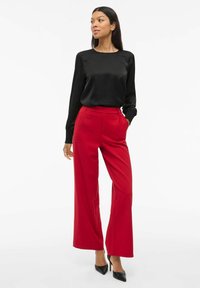 Blusa nera in satin a maniche lunghe abbinata a pantaloni larghi rossi a vita alta. Tacchi a punta nera completano l'aspetto dell'outfit.