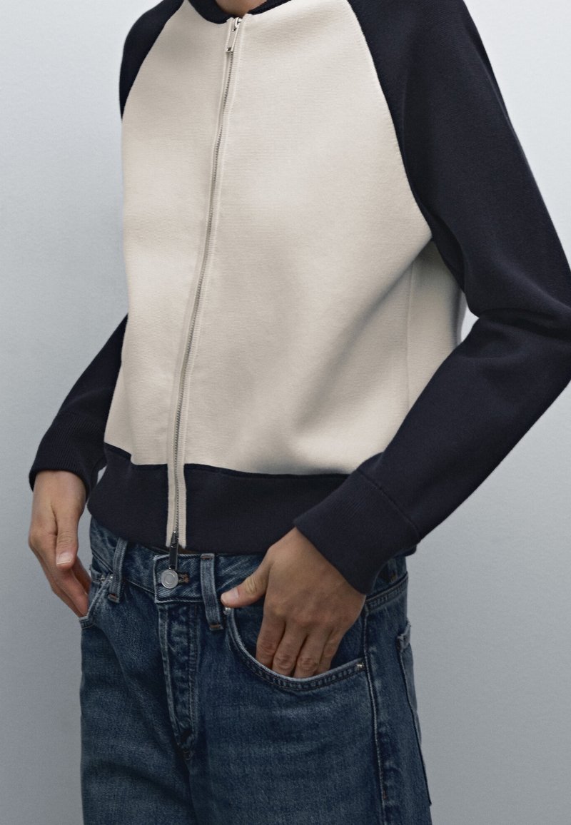 Personne portant une veste zippée noire et beige avec les mains dans les poches avant d'un jean en denim bleu.