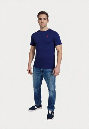 Fiatal férfi áll, rajta egy testhezálló sötétkék póló kis piros logóval, kék farmer és fekete-fehér sportcipő, egyszínű háttér előtt.