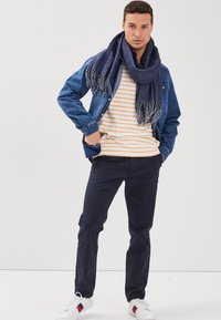 Jeansjacka, randig gul och vit t-shirt, marinblå byxor och en blå scarf med fransar. Vita sneakers med röda och blå detaljer.