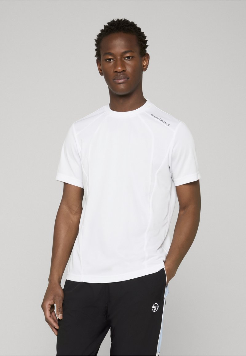 Jeune homme portant une chemise blanche à manches courtes et un pantalon noir avec des bandes latérales, debout devant un fond gris clair uni.