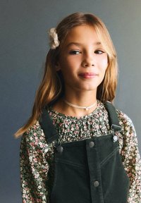 Dívka s dlouhými, světle hnědými vlasy, oblečená ve květinovém zeleném blůze pod tmavozeleným manšestrovým overalem s kovovými sponami a náhrdelníkem s chokerem.