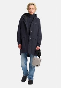 Manteau imperméable bleu marine avec capuche, grandes poches avant, boutons-pression, assorti à un jean bleu clair et un sac bandoulière gris.