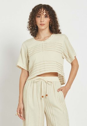 Ženska s kodrastimi lasmi nosi kremno teksturirano kratko majico z rokavi in široke hlače z vrvico v istem odtenku ter stoji z eno roko v žepu.
