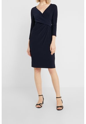 Vrouw draagt een marineblauwe wikkeljurk met lange mouwen en knielengte, met zwarte enkelbandhakken, staand tegen een effen lichte achtergrond.