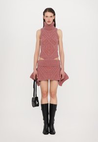 Set di top e gonna in maglia rosa a collo alto con motivi geometrici. Modella che tiene una borsa nera, indossando stivali neri fino al ginocchio.