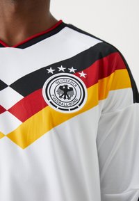 Λευκή αθλητική φανέλα με κόκκινες, μαύρες και κίτρινες διαγώνιες ρίγες; διαθέτει στρογγυλό λογότυπο με αετό με δύο κεφάλια και πέντε αστέρια.