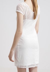 Bílé krajkové šaty s krátkými rolovanými rukávy, květinovou texturou, přiléhavým střihem a průhledným zadním panelem. Spodní lem sahá nad koleno.