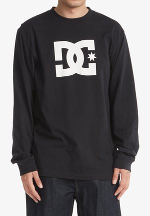 Personne portant un t-shirt noir à manches longues avec un grand logo blanc "DC" et une étoile centrée sur la poitrine, associé à un jean foncé.