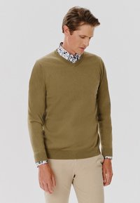 Oliwkowy sweter w serek wykonany z miękkiego materiału, z prążkowanym dołem i mankietami. Pod spodem widoczna jest biała koszula z czarnym wzorem kwiatowym.