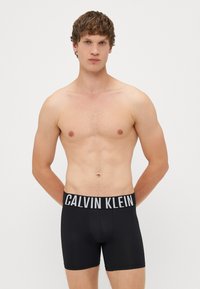 Boxeri scurți negri Calvin Klein cu un tiv elastic lat pe care apare numele brandului în alb. Material neted cu un design mulat.