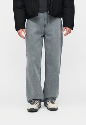 Άτομο που φοράει ξεθωριασμένα γκρι τζιν με φαρδιά μπατζάκια, μαύρο μπουφάν και λευκά αθλητικά παπούτσια με μαύρες σόλες σε απλό φόντο.