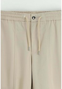 Pantalons de sport beiges avec une taille élastique, un cordon de serrage ajustable et des finitions en métal argenté. Texture lisse et coupe décontractée.