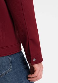 Burgundy jacka i texturerat tyg, med knapprad på manschetten och kontrasterande gul sömnad, bärs över blå denim.