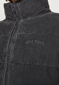 Grå kordflort, pufferjacka med vertikal ribbing, hög krage och dragkedjestängning. Har diskret broderi "BDG JEANS" på bröstet.