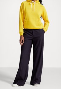 Gul hoodie med snörning och kenguruficka, kombinerad med högmidjade marinblå byxor och vita sneakers, som visar en avslappnad passform.
