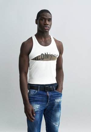 Jeune homme portant un débardeur blanc avec le texte découpé "ICONique" et un jean bleu usé, posant devant un fond gris uni.