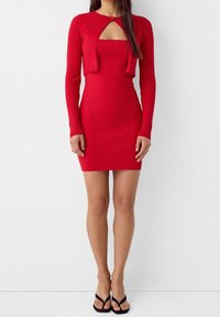 Czerwona sukienka z prążkowanej tkaniny z długimi rękawami, dekoltem w kształcie wycięcia i dopasowanym krojem. Materiał ma teksturowany wygląd. Zestawiona z czarnymi sandałami na cienkich paskach.