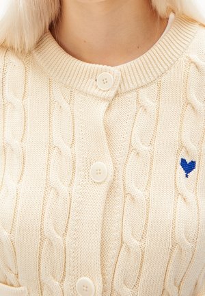 Cardigan crème en maille torsadée avec col rond, fermeture à boutons et petit cœur bleu brodé sur la poitrine.