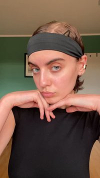 Fita de cabeça de tecido preto, design elegante, textura suave. Top preto, pele clara, brinco prateado. Mãos posicionadas sob o queixo, fundo neutro.