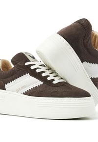 Braune Wildleder- und Mesh-Sneaker mit weißen Akzenten, ausgestattet mit einer dicken weißen Sohle, flachen Schnürsenkeln und einem Etikett auf der Zunge. Strukturierte Details sind vorhanden.