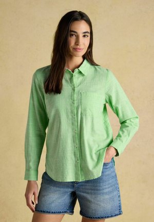 Kobieta o długich, ciemnych włosach, ubrana w jasną zieloną koszulę zapinaną na guziki i jeansowe szorty, stojąca z jedną ręką w kieszeni na beżowym tle.