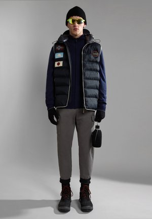 Mann trägt schwarze Mütze, neongelbe Sonnenbrille, schwarze gesteppte Weste mit Aufnähern, blaue Jacke, graue Cargohose, schwarze Handschuhe und Stiefel, hält eine schwarze Feldflasche.