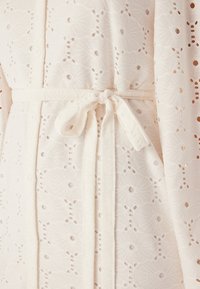 Witte stoffen robe met een ingewikkeld florale broderie anglaise patroon, vastgebonden met een bijpassende riem, die een zachte, textuurachtige uitstraling heeft.