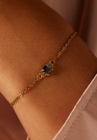 Gouden kettingarmband met een ovale zwarte steen als middelpunt, omringd door kleine heldere stenen, geplaatst tegen een textuur van huid.