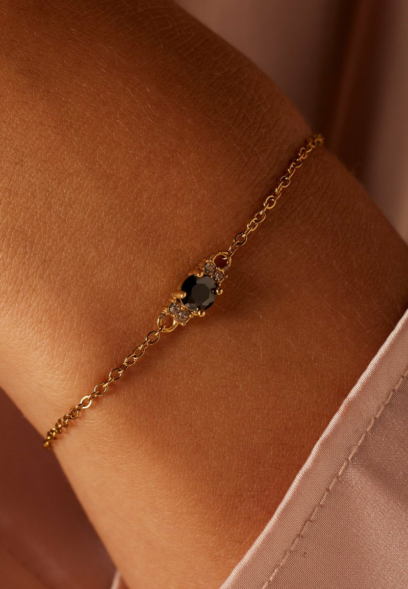 Gouden kettingarmband met een ovale zwarte steen als middelpunt, omringd door kleine heldere stenen, geplaatst tegen een textuur van huid.