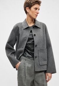 Γκρι σακάκι από μείγμα μαλλιού με γιακά, κλείσιμο με κουμπιά μπροστά και μεγάλη τσέπη. Συνδυασμένο με μαύρο τοπ και γκρι παντελόνι.