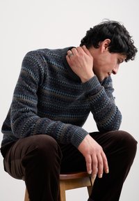 Vīrietis ar rakstainu džemperi un brūnām korduroja biksēm sēž uz koka krēsla, balstījis roku uz kakla un skatoties lejup.