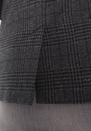 Gros plan sur un tissu à motif pied-de-poule noir et gris d'un blazer porté sur un pantalon gris clair, montrant un détail de fente latérale.