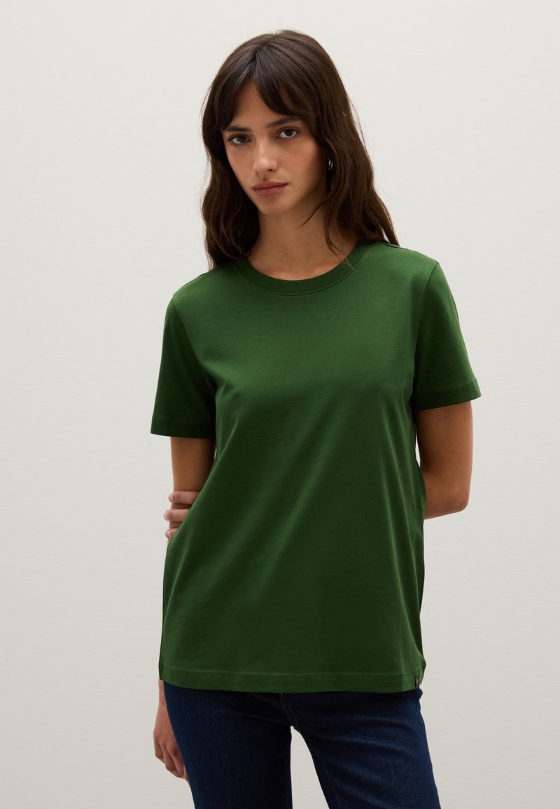 Grünes T-Shirt mit kurzen Ärmeln aus weichem Stoff, mit einem Rundhalsausschnitt, gerader Schnittführung und minimalen Details. Getragen mit blauen Jeans.