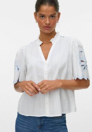 Blouse blanche à manches courtes avec un col à volants, accents brodés en bleu sur les manches, et un boutonnage sur le devant avec une coupe décontractée.