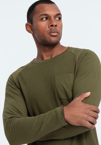 Oliivivihreä pitkähihainen paita, jossa on ribbitekstuuri ja rintatasku. Pehmeä kangas, pyöreä kaula-aukko ja istuvat hihat.