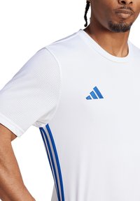 Camiseta atlética blanca con acentos azules, con una textura de malla en los hombros y tres franjas azules a lo largo del costado, y un logo azul.