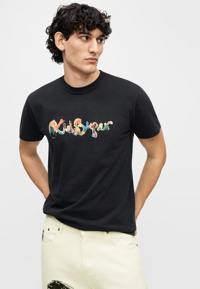 Μαύρο βαμβακερό μπλουζάκι με πολύχρωμη εκτύπωση γραφικού "KidSuper". Στρογγυλός λαιμός και κοντά μανίκια, φορεμένο με μπεζ παντελόνι.