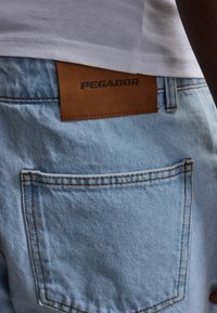Nahaufnahme einer Person, die hellblaue Jeans mit braunem Pegador-Label und ein weißes Hemd trägt, das leicht oberhalb des Bunds eingesteckt ist.