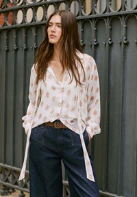 Camicia bianca floreale con maniche lunghe e laccetti in vita, abbinata a pantaloni di denim scuro a vita alta e una cintura marrone con fibbia argento.