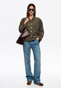 Oliwkowa kurtka zapinana na guziki z kieszenią, zestawiona z jasnoniebieskimi jeansami i brązowymi zamszowymi butami. Duże czarne okulary przeciwsłoneczne i brązowa torba.