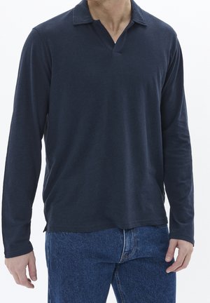 Man draagt een marineblauw overhemd met lange mouwen en kraag, en een blauwe spijkerbroek, staand tegen een effen witte achtergrond.