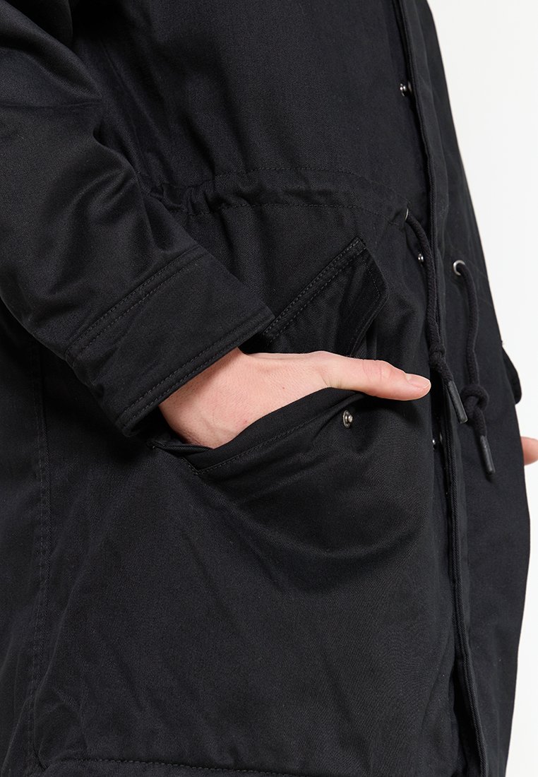 Veste extérieure noire avec une taille cintrée, des manches longues et des poches avant. Le matériau semble lisse avec une finition mate et un matériel minimal.