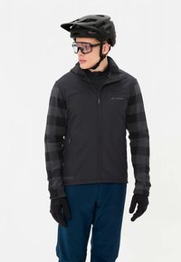 Zwarte jas met meerdere texturen, met grijze geruite mouwen, een ritssluiting aan de voorkant en zakken. Draagt een zwarte helm en doorzichtige bril, gecombineerd met een blauwe broek.