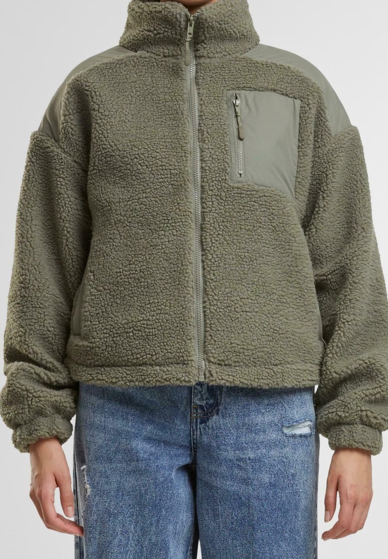 Fleece-Zip-Fleecejacke in Olivgrün mit hohem Kragen, Brusttasche mit Reißverschluss und strukturiertem Stoff; kombiniert mit zerrissenen blauen Jeans.