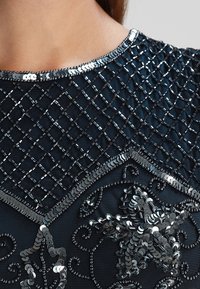 Svart tyg med intrikat pärlbroderi och paljetter. Mönstret inkluderar ett diamantmönster, accentuerat med glänsande, metalliska detaljer.