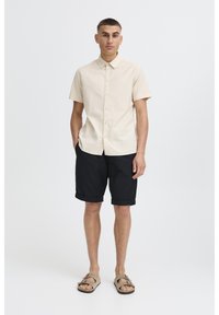 Short-sleeve beige shirt with button front, paired with black rolled shorts and tan sandals. Smooth fabric, relaxed fit, minimal design.